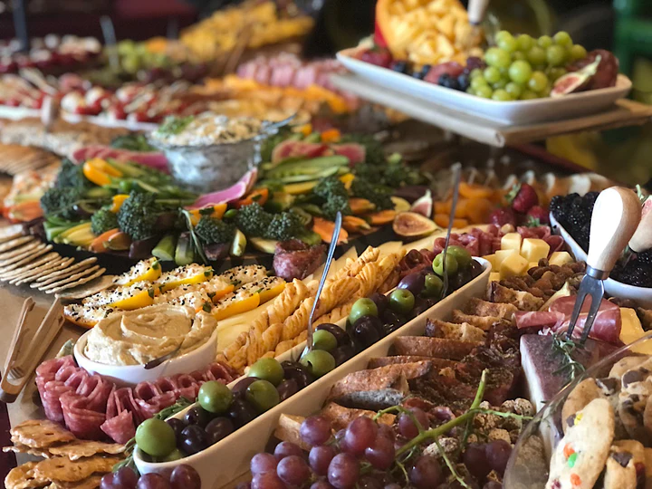 A picture of a buffet with catered food, such as grapes, olives, slices of bread, meet and veggie platters with dip